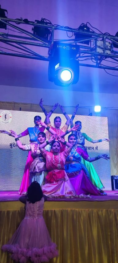 Five dancers in vibrant costumes performing on stage, with a child watching in awe.